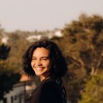 A joyful woman stands outdoors, smiling in the warm summer sun against a nature and urban backdrop.